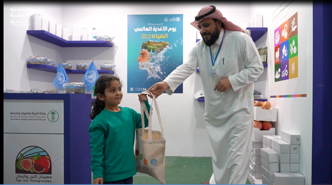 Photo of FAO staff member handing a Saudi young girl a goody bag from FAO booth at the event